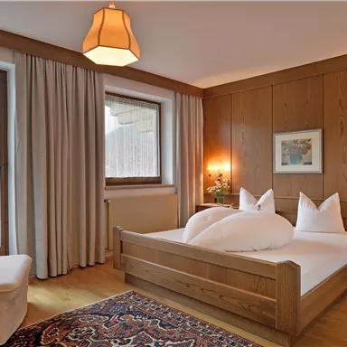 A cozy bedroom with wooden walls and a large window. The bed is adorned with white pillows and blankets, and a rug is laid on the floor.