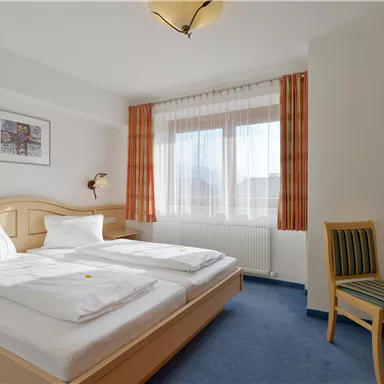 Ein helles Hotelzimmer mit einem Doppelbett und zwei Kissen. Neben dem Bett steht ein Stuhl und das Fenster bietet Blick auf den Außenbereich.