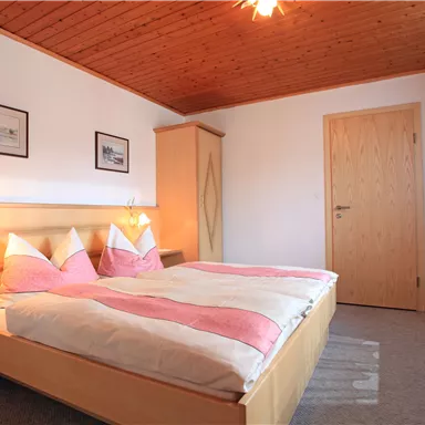 Ein gemütliches Schlafzimmer mit einem Doppelbett und rosa Bettwäsche. Die Wände sind hell, und es gibt einen Holzdecke mit einer warmen Beleuchtung.