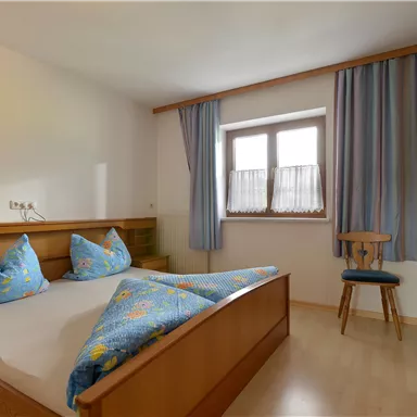 A simple bedroom with a double bed and two pillows. There is a chair and windows with curtains in a bright room.