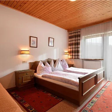 A cozy bedroom with two beds and a large window. The room is light and friendly decorated with wood paneling and carpet flooring.