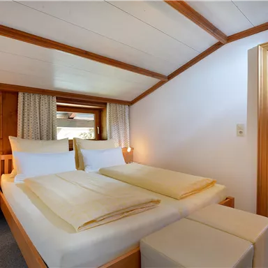 Ein gemütliches Schlafzimmer mit zwei Betten und warmen Holzelementen. Es gibt Fenster mit Vorhängen und eine angenehme Beleuchtung.