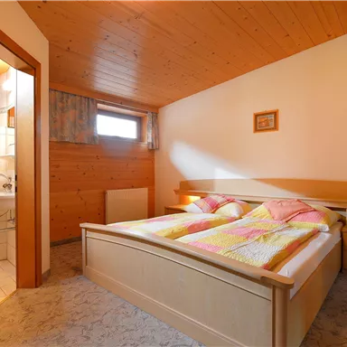 Ein gemütliches Schlafzimmer mit einem Doppelbett und Holzverkleidung. Im Hintergrund ist ein Badezimmer zu sehen.