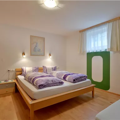 Ein gemütliches Schlafzimmer mit zwei Einzelbetten und bunten Kissen. Der Raum hat Holzfußboden und große Fenster, die für viel Tageslicht sorgen.