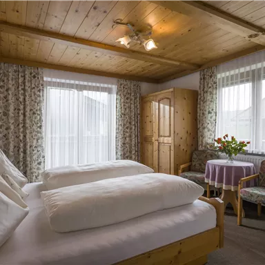 Ein gemütliches Schlafzimmer mit zwei Betten und Holzdecke. Durch die Fenster strömt Tageslicht, und ein Tisch mit Blumen sorgt für eine angenehme Atmosphäre.