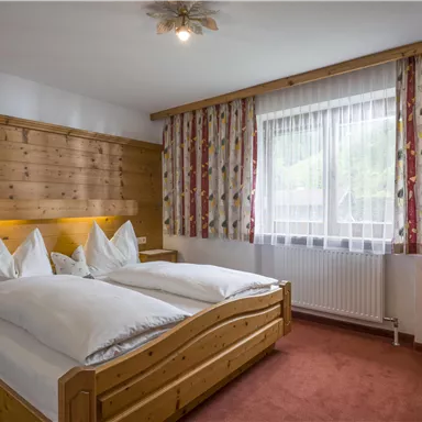 Ein gemütliches Schlafzimmer mit einem Doppelbett und großen Kissen. Die Wände sind aus hellem Holz, und es gibt Vorhänge mit buntem Muster.