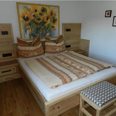 Ein gemütliches Schlafzimmer mit einem Doppelbett aus Holz und bunten Kissen. An der Wand hängt ein Gemälde mit Sonnenblumen.
