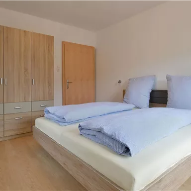 Ein gemütliches Schlafzimmer mit einem Doppelbett und blauen Bettdecken. Der Raum hat einen Kleiderschrank und eine Pflanze in einem Topf.