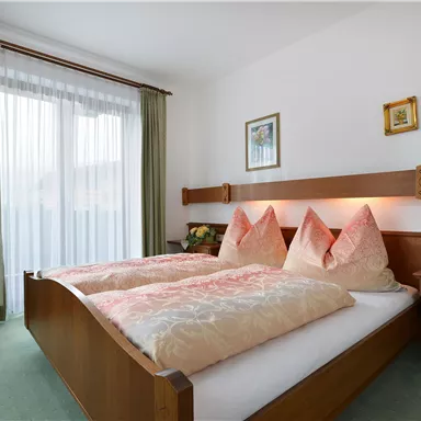 Ein gemütliches Schlafzimmer mit einem Doppelbett und zwei dekorativen Kissen. Helle Vorhänge lassen viel Tageslicht herein, und an der Wand hängen einige Bilder.