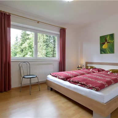 A cozy bedroom with a large bed and red bedcovers. The window offers a view of the nature outside.