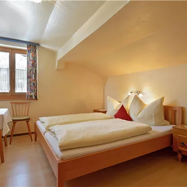 Ein gemütliches Zimmer mit einem Doppelbett und hellem Holzinterieur. Es gibt ein Fenster mit Vorhängen und einen kleinen Tisch mit einem Stuhl.