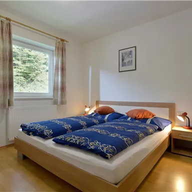 A cozy bedroom with a large bed and blue covers. The walls are light and a window provides daylight.