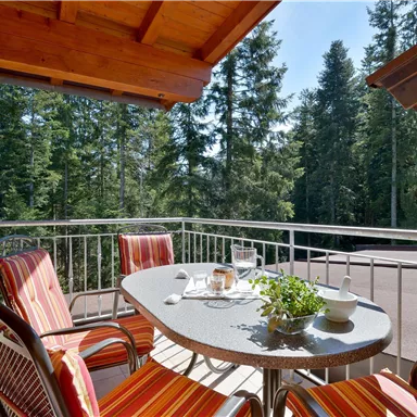 Ein gemütlicher Balkon mit einem Tisch und zwei Stühlen, umgeben von grünen Bäumen. Auf dem Tisch stehen Kaffee und ein kleines Pflänzchen.