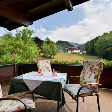 Ein einladender Balkon mit gemütlichen Stühlen und einem Tisch, auf dem Getränke stehen. Im Hintergrund sind grüne Wiesen und Bäume sowie sanfte Hügel zu sehen.
