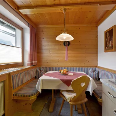 A cozy kitchen with wooden paneling and a round table. On the table, there is a bowl of fruit and the windows are decorated with curtains.