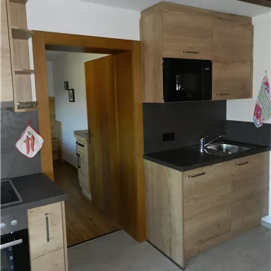 Eine moderne Küche mit Holzschränken und grauen Arbeitsflächen. Im Hintergrund führt eine Tür weiter in den Raum.