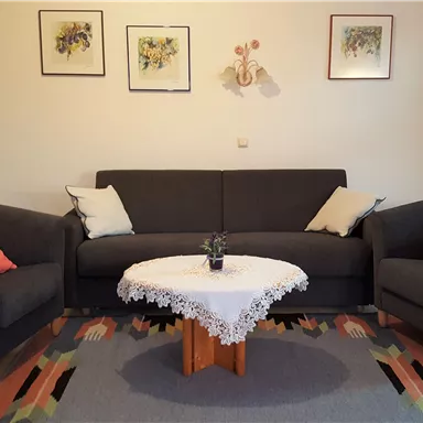 Ein gemütliches Wohnzimmer mit einem grauen Sofa und zwei Sessel. Tisch mit einer weißen Tischdecke und einer Pflanze in der Mitte.
