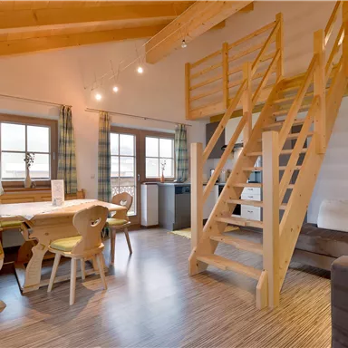 Ein gemütliches Wohnzimmer mit Holzdecken und großen Fenstern. Eine Treppe führt zu einem oberen Bereich, und es gibt Ess- und Sitzgelegenheiten.