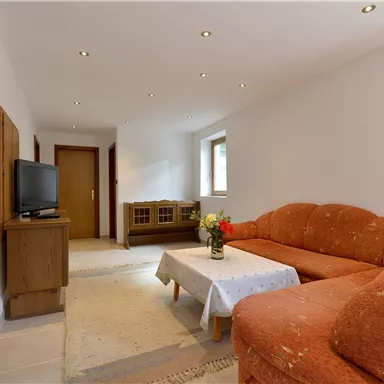A bright living room with an orange sofa and a table in the middle. There is a television and a display case on the wall.
