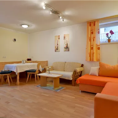 Ein gemütliches Wohnzimmer mit orangefarbenen Sofas und einem Esstisch. Warme Farben und Tageslicht schaffen eine einladende Atmosphäre.