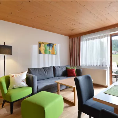 Ein modernes Wohnzimmer mit einem grauen Sofa und einem grünen Sessel. Große Fenster bieten Blick auf die Natur draußen.