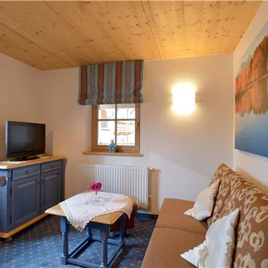 A cozy living space with a sofa and a small table. A picture with a landscape motif hangs on the wall, and a television stands in the corner.