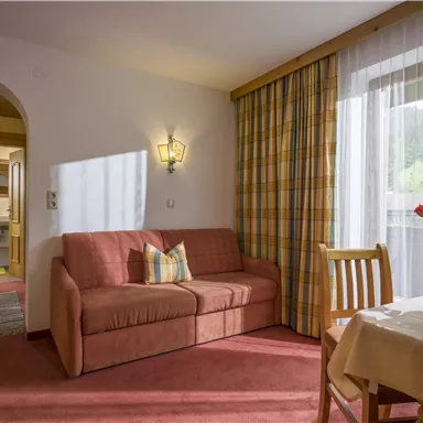 Eine gemütliche Wohnstube mit einem Sofa und einem Essbereich. Die Fenster lassen viel Licht herein und bieten einen Blick auf die Umgebung.