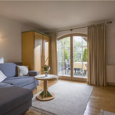 Ein gemütliches Wohnzimmer mit einem grauen Sofa und einem kleinen Tisch. Große Fenster bieten Blick auf die Terrasse und viel Tageslicht.