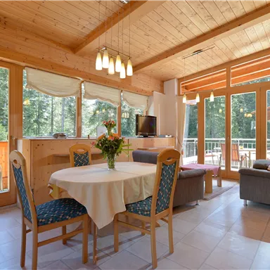 Helles und modernes Wohnzimmer mit großen Fenstern und Blick ins Grüne. Ein runder Esstisch und bequeme Sitzgelegenheiten laden zum Entspannen ein.