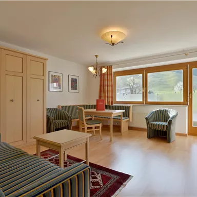 Ein gemütliches Wohnzimmer mit einem Sofa und zwei Sesseln. Große Fenster lassen viel Licht herein und bieten einen Blick nach draußen.