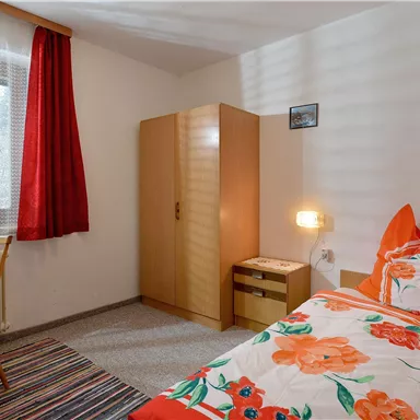 Ein gemütliches Einzelzimmer mit einem Bett und bunten Blumenbettwäsche. Der Raum verfügt über einen Schreibtisch, einen Stuhl und einen Kleiderschrank.
