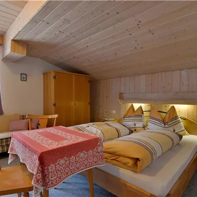 Ein gemütliches Zimmer mit einem Doppelbett, Holzverkleidung und sanfter Beleuchtung. Ein kleiner Tisch mit einer Tischdecke und Stühlen vervollständigt das Ambiente.