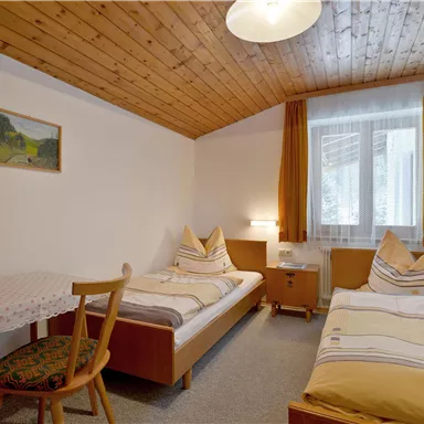 Ein gemütliches Schlafzimmer mit zwei Betten, einem Tisch und einer hellen Fensterfront. Die Wände sind in neutralen Farben gehalten, und es gibt eine Holzdecke für ein warmes Ambiente.
