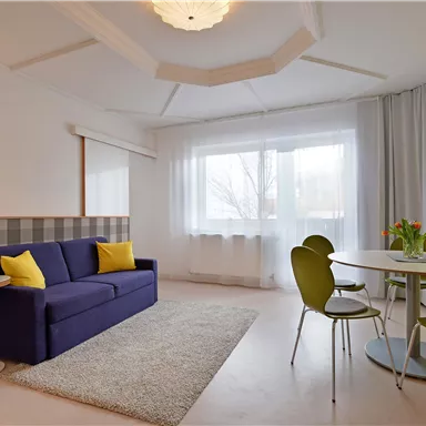 Ein helles und modernes Wohnzimmer mit einem blauen Sofa und einem Esstisch. Die großen Fenster sorgen für viel natürliches Licht.