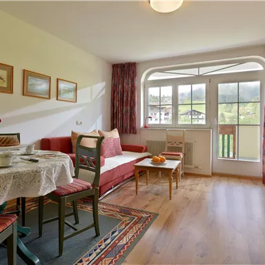 Ein gemütliches Wohnzimmer mit einem Essbereich und großen Fenstern. Die Möbel sind in warmen Farben gehalten, und ein kleiner Balkon ist sichtbar.