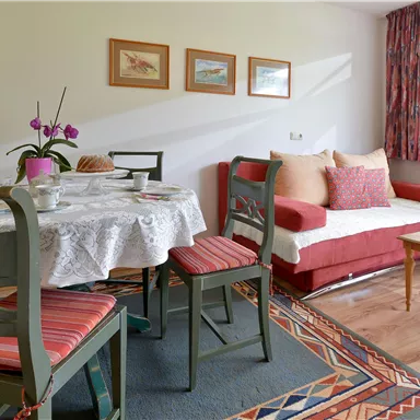 Ein gemütlicher Wohnraum mit einem Esstisch und Stühlen, einer roten Couch und bunten Vorhängen. Auf dem Tisch liegen frisches Obst und eine Tischdecke mit Blumenmuster.