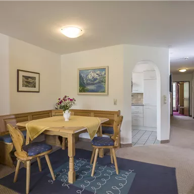 Ein gemütlicher Essbereich mit einem Holztisch und Stühlen. Im Hintergrund ist eine offene Küche und ein Bild an der Wand zu sehen.