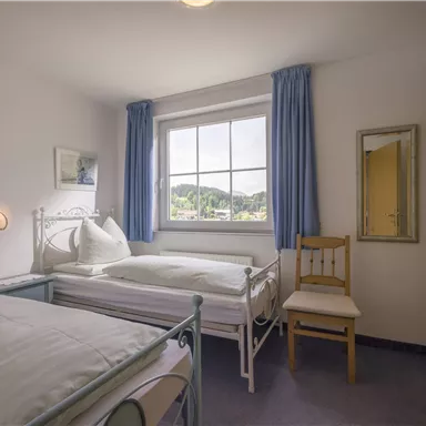 Ein helles Schlafzimmer mit zwei Einzelbetten und einem großen Fenster. Der Raum ist schlicht eingerichtet mit einer Kommode und einem Stuhl.