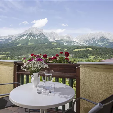 Eine Terrasse mit einem kleinen Tisch und zwei Stühlen blickt auf eine atemberaubende Berglandschaft. Bunte Blumen schmücken das Geländer und die Umgebung ist klar und sonnig.