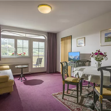 Ein gemütlicher Wohnraum mit einem Sofa, einem Essbereich und einem Fenster, das auf eine grüne Landschaft blickt. Die Dekoration ist einfach und einladend.