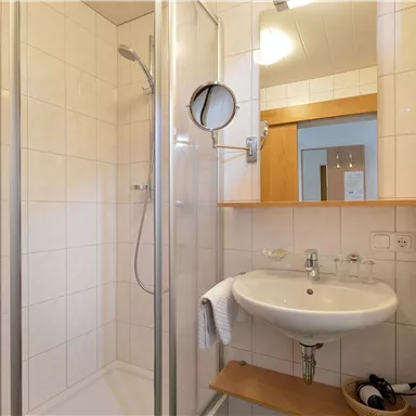 Ein modernes Badezimmer mit einer Dusche und einem Waschbecken. Die Wände sind hell gefliest und es gibt eine Holzablage.