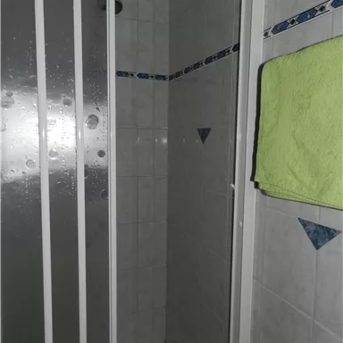 Eine moderne Duschkabine mit grauen Fliesen und einer grünen Handtuch. Die Tür ist aus transparentem Glas und es gibt Wasserflecken auf der Glasoberfläche.