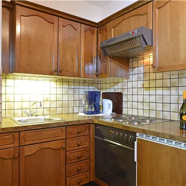 Eine moderne Küche mit Holzschränken, Waschbecken und Küchengeräten. Auf der Arbeitsplatte stehen ein Wasserkocher, eine Flasche Wein und Gläser.