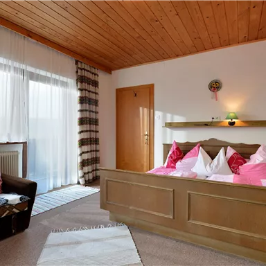 A cozy bedroom with a double bed and pink pillows. The room has large windows with curtains and a modern seating area.