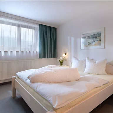 A bright bedroom with a large double bed and two pillows. The walls are painted in neutral colors, and there are curtains as well as a window that lets in daylight.