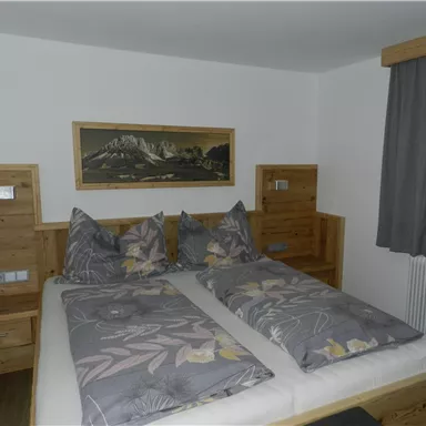 Ein gemütliches Schlafzimmer mit einem Doppelbett und Holzmöbeln. Die Wände sind hell gestrichen und ein Kunstwerk mit Bergmotiven hängt über dem Bett.