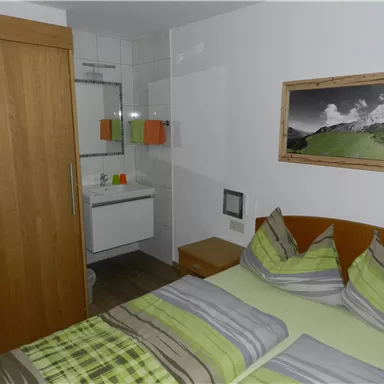 Ein gemütliches Zimmer mit einem Doppelbett und bunten Kissen. Im Hintergrund sind ein Badezimmer und ein Schrank sichtbar.