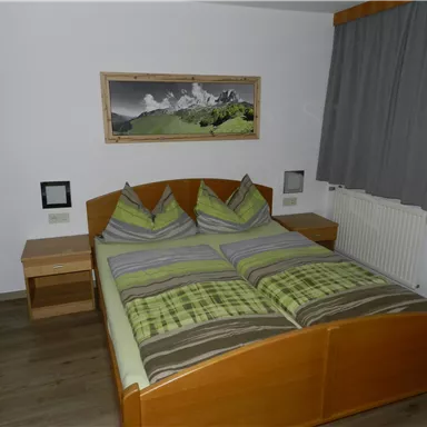 Ein einfaches Schlafzimmer mit einem Doppelbett und holzfarbener Einrichtung. An der Wand hängt ein Bild von Bergen, und die Fenster sind mit Vorhängen dekoriert.