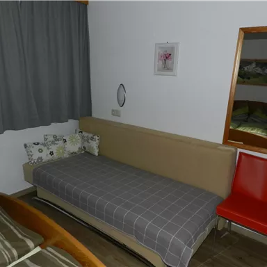 Ein schlichtes Zimmer mit einem Bett, einem roten Stuhl und einem Spiegel an der Wand. Der Raum hat eine graue Gardine und eine gemütliche Atmosphäre.