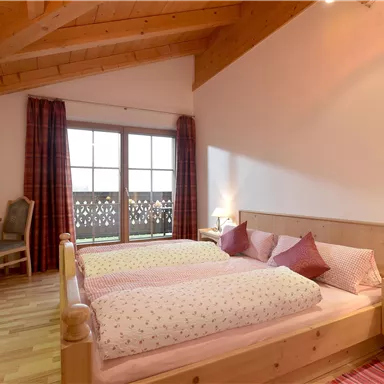 Ein gemütliches Schlafzimmer mit einem Doppelbett und Holzmöbeln. Große Fenster sorgen für viel Tageslicht und einen Blick auf den Balkon.
